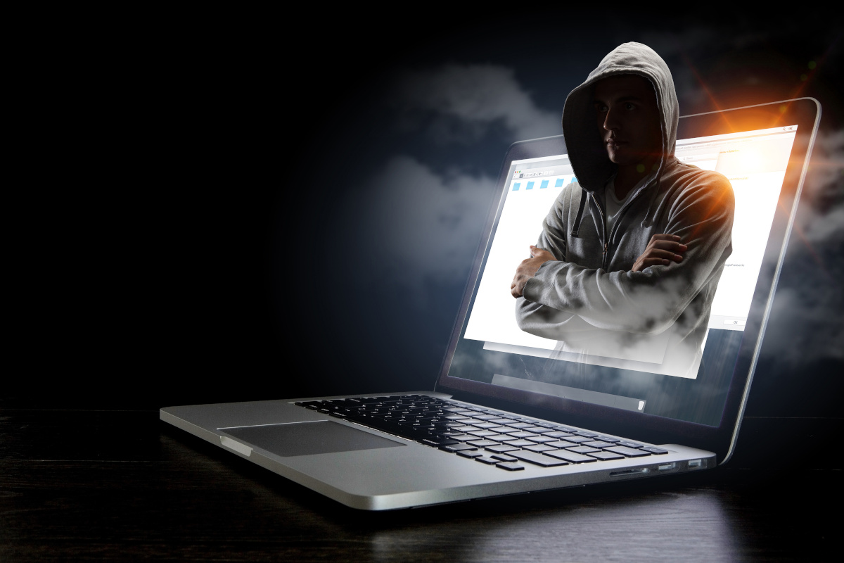 A person in a hoodie appearing out of a laptop screen in a dark room