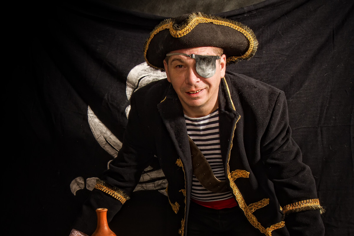 An adult pirate in a cocked hat and a striped vest plays cards on a wooden table against the background of the Jolly Roger flag.