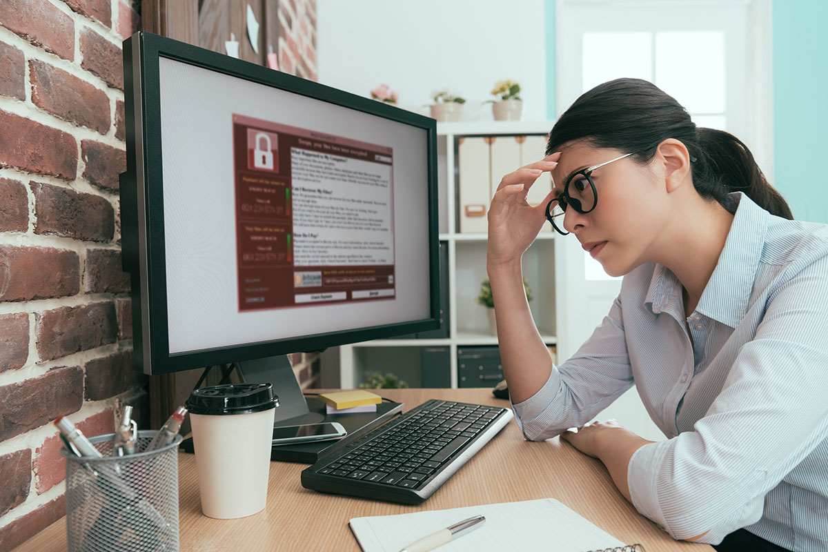 Frustrated business woman holding head at desk with computer screen showing security alert.